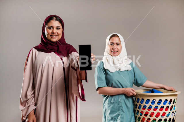 منصات إلكترونية لاستقدام العماله المنزليه المدربة، الخدمات الإلكترونيه المتقدمه لدعم وتوفير الخادمات المرخصة والمؤهله، بورتريه لسيدة محجبة تحمل بيدها هاتف بشاشة سوداء فارغة وعاملة فلبينية تحمل بيدها سلة الملابس، مهن ووظائف نسائية، خلفية رمادية