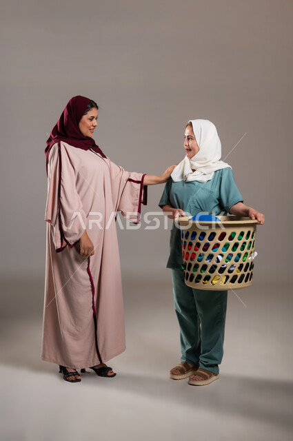 Routine household tasks and chores, a portrait of a veiled Saudi Gulf Arab woman supervising and directing the housekeeper at the laundry service, a Filipino woman wearing a work uniform and carrying a basket of clothes, women’s professions and jobs, full length of the body, gray background