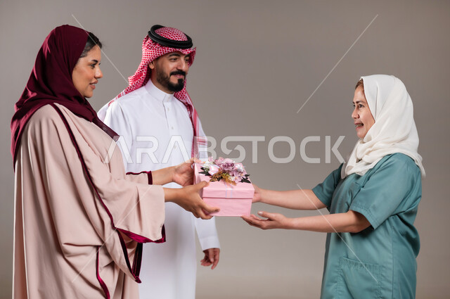 الإحسان في التعامل مع الخادمات في البيوت السعودية، القيام بالمهام والأعمال المنزلية الروتينية، بورتريه لرجل وامرأة عربيان سعوديان خليجيان يقومان بتقديم هديه للعاملة الفلبينية بايماءات الشكر والسرور لجودة خدمتها وادائها، مهن ووظائف نسائية، خلفية رمادية