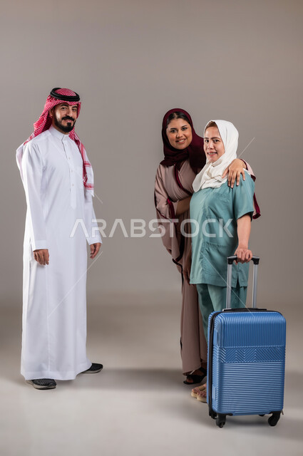 المعاملة الحسنة للخادمات، تأجيرالعمالة الخارجية المؤهلة من الخارج، بورتريه لرجل وامرأة عربيان سعوديان خليجيان يستقبلان عاملة فلبينية ترتدي اليونيفورم الخاص بالعمل وتمسك بيدها حقيبة السفر، النظر الى الكاميرا بايماءات السرور، الطول الكامل للجسم, خلفية رمادية