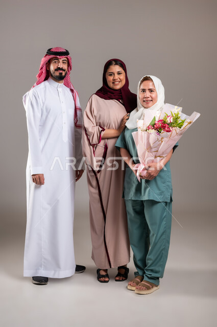 النظر الى الكاميرا بايماءات الشكر والامتنان لجودة خدمة واداء العاملة المنزلية، بورتريه لرجل وامرأة عربيان سعوديان خليجيان يقدمان باقة زهور الى العاملة الفلبينية، استقدام العمالة الخارجية المرخصة والمؤهلة، المعاملة الحسنة للخادمات، الطول الكامل للجسم، خلفية رمادية