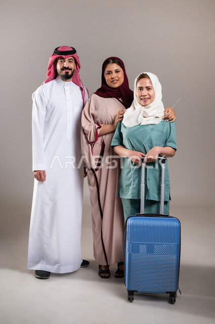 النظر الى الكاميرا بايماءات السرور، المعاملة الحسنة للخادمات، تأجيرالعمالة الخارجية المؤهلة من الخارج، بورتريه لرجل وامرأة عربيان سعوديان خليجيان يستقبلان عاملة فلبينية ترتدي اليونيفورم الخاص بالعمل وتمسك بيدها حقيبة السفر، الطول الكامل للجسم, خلفية رمادية