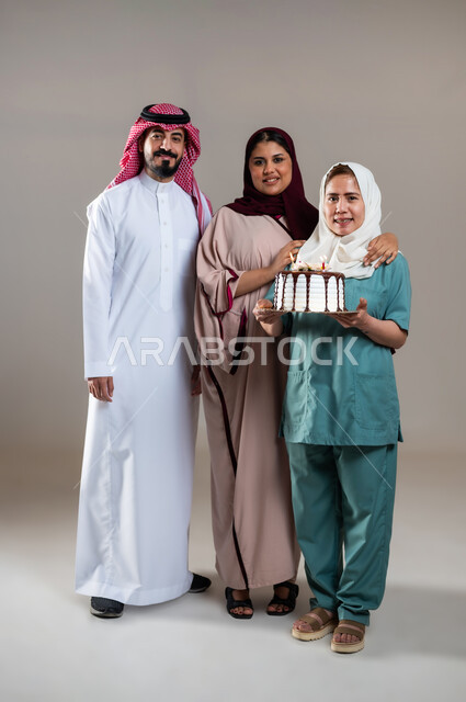 النظر الى الكاميرا بايماءات السعادة و السرور, بورتريه لزوجان عربيان سعوديان خليجيان يحتفلان بالعاملة الفيلبينية بالكيك المزين بالشموع،  قضاء أوقات ممتعه بالمناسبات وأعياد الميلاد, الإحسان في التعامل مع الخادمات في البيوت السعودية، الطول الكامل للجسم، خلفية رمادية