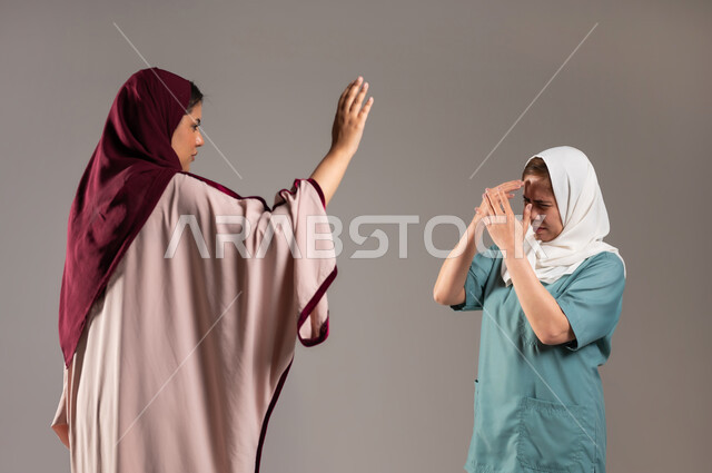 Gestures of shouting, reprimanding, and blaming, a portrait of a Saudi Gulf Arab woman raising her hands in annoyance to hit the Filipino maid for not completing the work as required, the worker walking away and crying with gestures of fear and panic, gray background.
