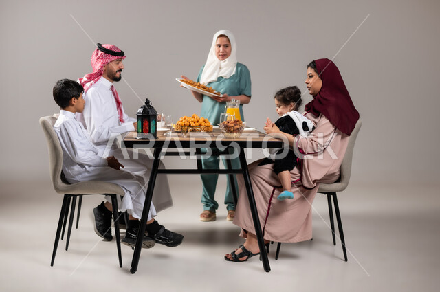 Islamic religious atmosphere and worship, a family Ramadan breakfast, an iftar table full of delicious food and desserts, a portrait of a Saudi Gulf Arab family sitting on a dining table before breakfast in the holy month of Ramadan, attracting workers from abroad, gray background