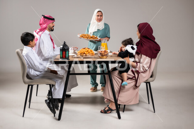 أجواء وعبادات دينية إسلامية، فطور رمضاني عائلي, مائدة إفطار مليئة بأصناف المأكولات والحلويات الشهية, بورتريه لأسرة عربية خليجية سعودية تجلس على سفرة الطعام  قبل موعد الإفطار في شهر رمضان المبارك، استقطاب العمال من الخارج، خلفية رمادية