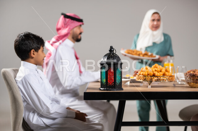 فانوس رمضاني تقليدي ملون ،أجواء وعبادات دينية إسلامية، فطور رمضاني عائلي, مائدة إفطار مليئة بأصناف المأكولات والحلويات الشهية, أسرة عربية خليجية سعودية تجلس على سفرة الطعام  قبل موعد الإفطار في شهر رمضان المبارك، استقطاب العمال من الخارج، خلفية رمادية