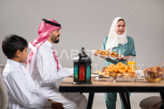 فانوس رمضاني تقليدي ملون ،أجواء وعبادات دينية إسلامية، فطور رمضاني عائلي, مائدة إفطار مليئة بأصناف المأكولات والحلويات الشهية, أسرة عربية خليجية سعودية تجلس على سفرة الطعام  قبل موعد الإفطار في شهر رمضان المبارك، استقطاب العمال من الخارج، خلفية رمادية