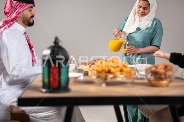 استقطاب عمال من الخارج، أجواء وطقوس دينية إسلامية، إفطار رمضاني عائلي، مائدة إفطار مليئة بالمأكولات اللذيذة والحلويات، عائلة عربية خليجية سعودية تجلس على طاولة طعام قبل الإفطار في شهر رمضان المبارك، خلفية رمادية.
