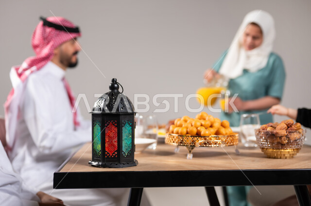 أسرة عربية خليجية سعودية تجلس على سفرة الطعام  قبل موعد الإفطار في شهر رمضان المبارك، أجواء وعبادات دينية إسلامية، فطور رمضاني عائلي, مائدة إفطار مليئة بأصناف المأكولات والحلويات الشهية، استقطاب العمال من الخارج، خلفية رمادية