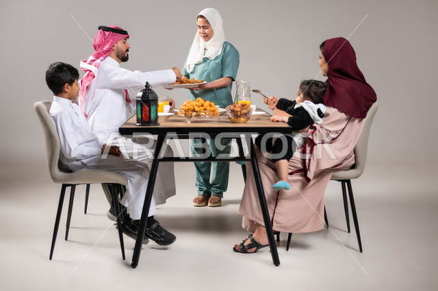 مائدة إفطار مليئة بأصناف المأكولات والحلويات الشهية، أجواء وعبادات دينية إسلامية، فطور رمضاني عائلي, أسرة عربية خليجية سعودية تجلس على سفرة الطعام  قبل موعد الإفطار في شهر رمضان المبارك، استقطاب العمال من الخارج، خلفية رمادية