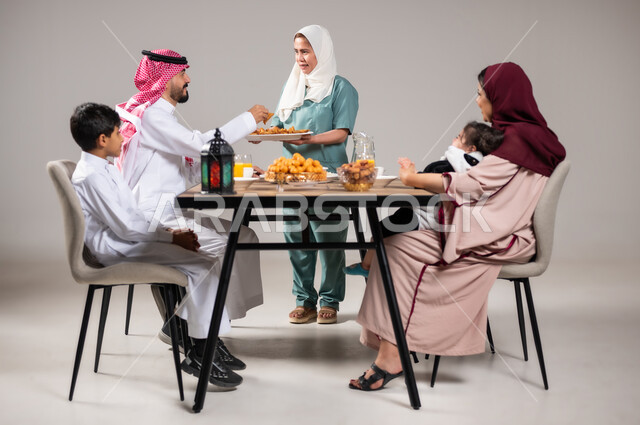 فطور رمضاني عائلي، أجواء وعبادات دينية إسلامية, مائدة إفطار مليئة بأصناف المأكولات والحلويات الشهية, أسرة عربية خليجية سعودية تجلس على سفرة الطعام  قبل موعد الإفطار في شهر رمضان المبارك، استقطاب العمال من الخارج، خلفية رمادية