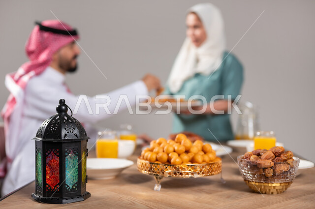 وجبة افطار ذات قيمة غذائية عالية, كرات العجين المقلية الصغيرة, مأكولات صحية على المائدة في شهر رمضان, بورتريه مقرب لعاملة منزلية تقوم بتقديم الحلويات لرجل عربي خليجي سعودي، طبق مليء بحبات من التمر الطازج بجانب فانوس ملون، حلا الزلابيات الشرقي، خلفية رمادية