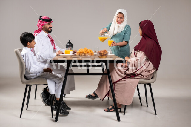 ايماءات السعادة بقدوم الشهر الفضيل,عائلة عربية خليجية سعودية مجتمعة على طاولة الطعام، الاحتفال بالمناسبات و الاعياد الاسلامية, العصائر و الاصناف الشرقية اللذيذة, بورتريه مقرب لعاملة منزلية تقوم بصب العصير و توزيعه, تنظيم سفرة فطور رمضان, خلفية رمادية