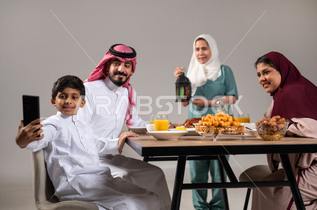 فطور رمضاني عائلي, مائدة إفطار مليئة بأصناف المأكولات الشهية, أسرة عربية خليجية سعودية تجلس على سفرة الطعام  قبل موعد الإفطار في شهر رمضان المبارك, عاملة فلبينية تحمل الفانوس الملون، تعظيم وتقدير الشهر الفضيل، خلفية رمادية