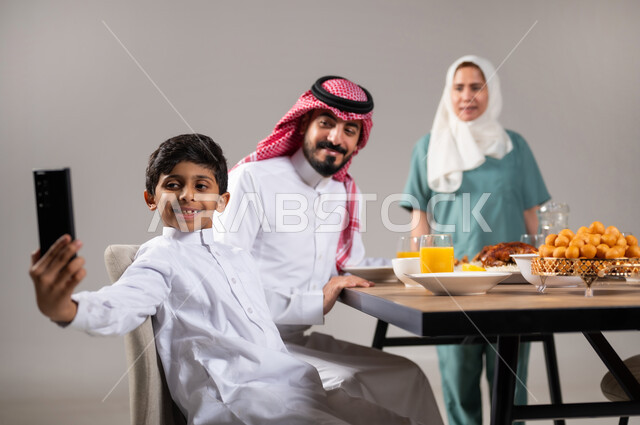 التقاط صور تذكارية، قضاء أوقات ممتعة مع العائلة, تحضير وجبة الإفطار، ايماءات وتعابير تدل على السعادة, أجواء عائلية سعيدة, بورتريه لعاملة فلبينية وعائلة عربية خليجية سعودية تجلس على مائدة الطعام المليئة بأصناف متنوعة وشهية في شهر رمضان المبارك، خلفية رمادية