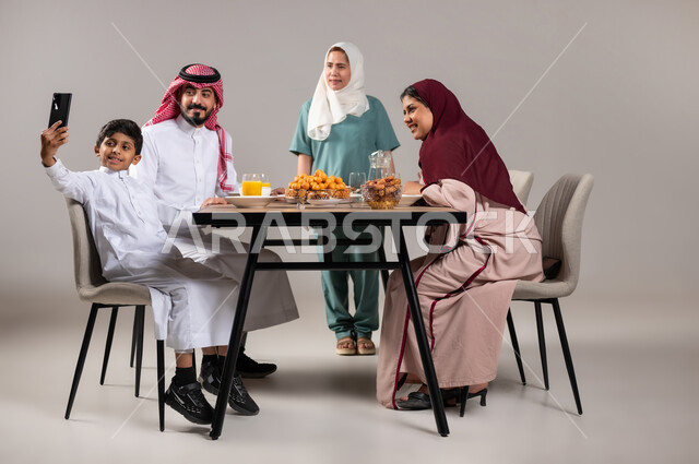 التقاط صور تذكارية، قضاء أوقات ممتعة مع العائلة, تحضير وجبة الإفطار، ايماءات وتعابير تدل على السعادة, أجواء عائلية سعيدة, بورتريه لعاملة فلبينية وعائلة عربية خليجية سعودية تجلس على مائدة الطعام المليئة بأصناف متنوعة وشهية في شهر رمضان المبارك، خلفية رمادية