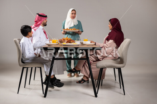A happy family atmosphere, a portrait of a Filipina worker preparing a Ramadan dinner and placing the main dish on it, appetizers and oriental Arabic dishes, gestures of happiness on the occasion of the advent of the holy month, celebrating Islamic holidays and occasions, family gathering at the dining table, gray background