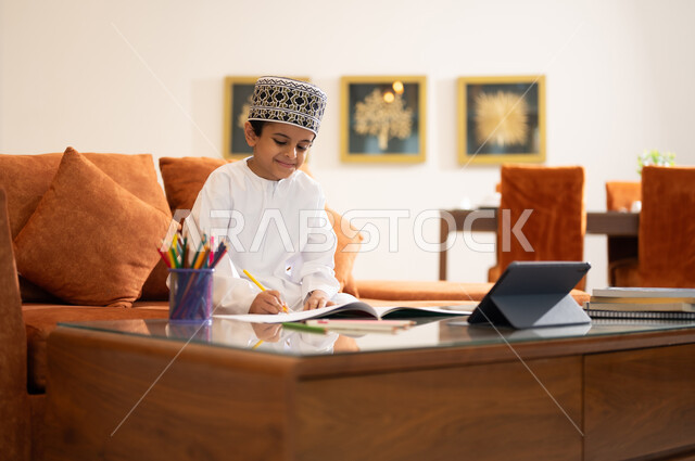 مراجعة الدروس اليومية عبر الانترنت، تطور العلم والتعليم في سلطنه عمان، بورتريه لتلميذ عربي خليجي عماني يرتدي الدشداشه والكمه يجلس في غرفه المعيشه امام جهاز التابلت المحمول يقوم بحل الواجبات المدرسيه وتلقي المحاضرات المدرسيه عن بعد