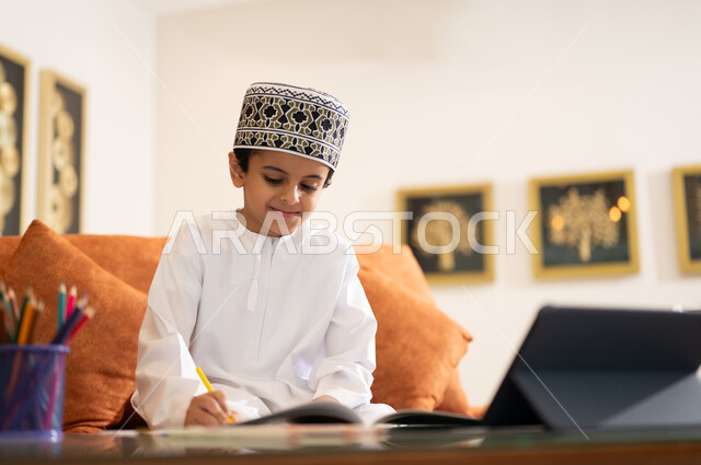 مراجعة الدروس اليومية عبر الانترنت، تطور العلم والتعليم في سلطنه عمان، بورتريه لتلميذ عربي خليجي عماني يرتدي الدشداشه والكمه يجلس في غرفه المعيشه امام جهاز التابلت المحمول يقوم بحل الواجبات المدرسيه وتلقي المحاضرات المدرسيه عن بعد