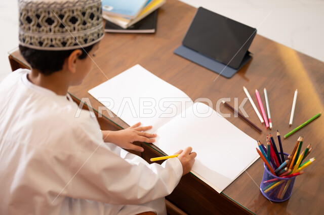 مراجعة الدروس اليومية عبر الانترنت، تطور العلم والتعليم في سلطنه عمان، بورتريه لتلميذ عربي خليجي عماني يرتدي الدشداشه والكمه يجلس في غرفه المعيشه امام جهاز التابلت المحمول يقوم بحل الواجبات المدرسيه وتلقي المحاضرات المدرسيه عن بعد