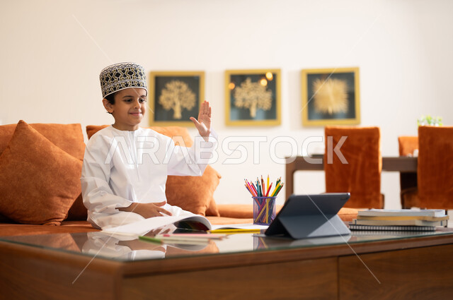 مراجعة الدروس اليومية عبر الانترنت، تطور العلم والتعليم في سلطنه عمان، بورتريه لتلميذ عربي خليجي عماني يرتدي الدشداشه والكمه يجلس في غرفه المعيشه امام جهاز التابلت المحمول يقوم بحل الواجبات المدرسيه وتلقي المحاضرات المدرسيه عن بعد يشير بيده نحو الاعلى