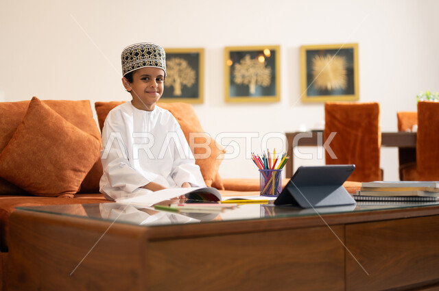 مراجعة الدروس اليومية عبر الانترنت، تطور العلم والتعليم في سلطنه عمان، بورتريه لتلميذ عربي خليجي عماني يرتدي الدشداشه والكمه يجلس في غرفه المعيشه امام جهاز التابلت المحمول يقوم بحل الواجبات المدرسيه وتلقي المحاضرات المدرسيه عن بعد