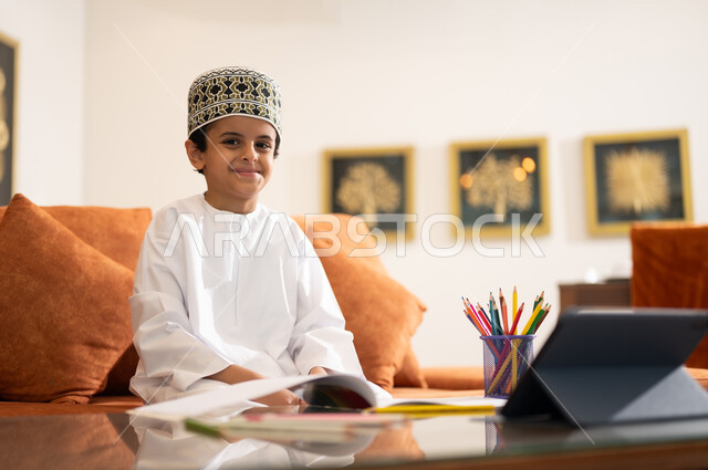 مراجعة الدروس اليومية عبر الانترنت، تطور العلم والتعليم في سلطنه عمان، بورتريه لتلميذ عربي خليجي عماني يرتدي الدشداشه والكمه يجلس في غرفه المعيشه امام جهاز التابلت المحمول يقوم بحل الواجبات المدرسيه وتلقي المحاضرات المدرسيه عن بعد