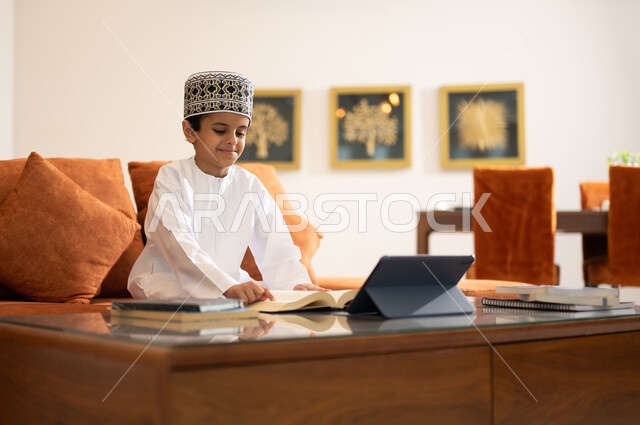 مراجعة الدروس اليومية عبر الانترنت، تطور العلم والتعليم في سلطنه عمان، بورتريه لتلميذ عربي خليجي عماني يرتدي الدشداشه والكمه يجلس في غرفه المعيشه امام جهاز التابلت المحمول يقوم بحل الواجبات المدرسيه وتلقي المحاضرات المدرسيه عن بعد