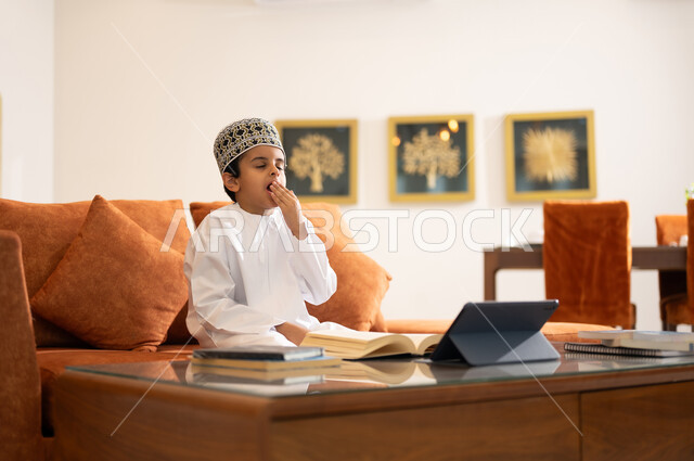 مراجعة الدروس اليومية عبر الانترنت، تطور العلم والتعليم في سلطنه عمان، بورتريه لتلميذ عربي خليجي عماني يرتدي الدشداشه والكمه يجلس في غرفه المعيشه امام جهاز التابلت المحمول يقوم بحل الواجبات المدرسيه وتلقي المحاضرات المدرسيه عن بعد