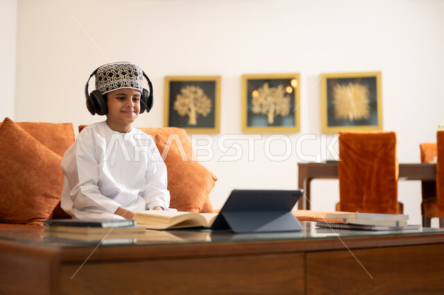 مراجعة الدروس اليومية عبر الانترنت، تطور العلم والتعليم في سلطنه عمان، بورتريه لتلميذ عربي خليجي عماني يرتدي الدشداشه والكمه يجلس في غرفه المعيشه امام جهاز التابلت المحمول يقوم بحل الواجبات المدرسيه وتلقي المحاضرات المدرسيه عن بعد