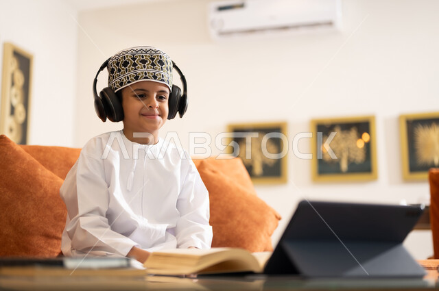 مراجعة الدروس اليومية عبر الانترنت، تطور العلم والتعليم في سلطنه عمان، بورتريه لتلميذ عربي خليجي عماني يرتدي الدشداشه والكمه يجلس في غرفه المعيشه امام جهاز التابلت المحمول يقوم بحل الواجبات المدرسيه وتلقي المحاضرات المدرسيه عن بعد