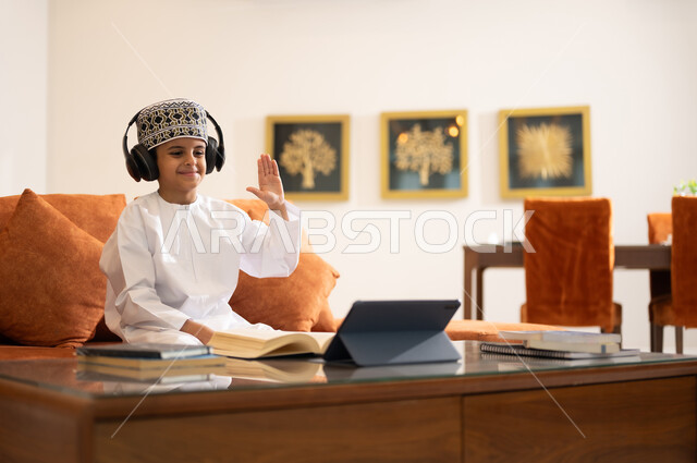 مراجعة الدروس اليومية عبر الانترنت، تطور العلم والتعليم في سلطنه عمان، بورتريه لتلميذ عربي خليجي عماني يرتدي الدشداشه والكمه يجلس في غرفه المعيشه امام جهاز التابلت المحمول يرتدي جهاز الهيدفون يستمع للدروس يقوم بحل الواجبات المدرسيه وتلقي المحاضرات المدرسيه عن بعد