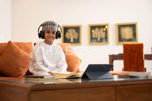 مراجعة الدروس اليومية عبر الانترنت، تطور العلم والتعليم في سلطنه عمان، بورتريه لتلميذ عربي خليجي عماني يرتدي الدشداشه والكمه يجلس في غرفه المعيشه امام جهاز التابلت المحمول يرتدي جهاز الهيدفون يستمع للدروس يقوم بحل الواجبات المدرسيه وتلقي المحاضرات المدرسيه عن بعد