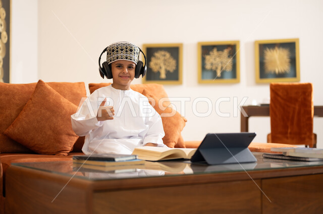مراجعة الدروس اليومية عبر الانترنت، تطور العلم والتعليم في سلطنه عمان، بورتريه لتلميذ عربي خليجي عماني يرتدي الدشداشه والكمه يجلس في غرفه المعيشه امام جهاز التابلت المحمول يرتدي جهاز الهيدفون يستمع للدروس يقوم بحل الواجبات المدرسيه وتلقي المحاضرات المدرسيه عن بعد