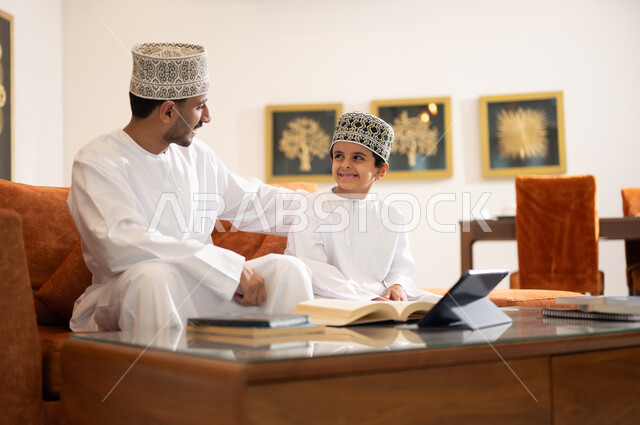 تنميه قدرات الاطفال بالتعلم، تشجيع وتطوير الاطفال بحل الواجبات المدرسيه المنزليه، بورتريه لأب عربي خليجي عماني يرتدي الدشداشه والكمه يقوم بمساعده ابنه في التعليم والقراءه، الانشطة التعليمية الالكترونيه  وانجاز الدروس والواجبات عن بعد باستخدام جهاز الايباد المحمول