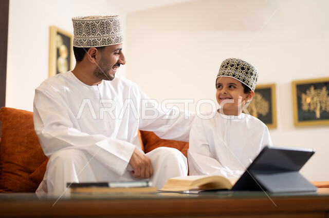Developing children’s learning abilities, encouraging and developing children by solving home school assignments, a portrait of an Arab, Gulf, Omani father wearing a dishdasha and a sleeve, helping his son in education and reading, electronic educational activities, and completing lessons and assignments remotely using a portable iPad.