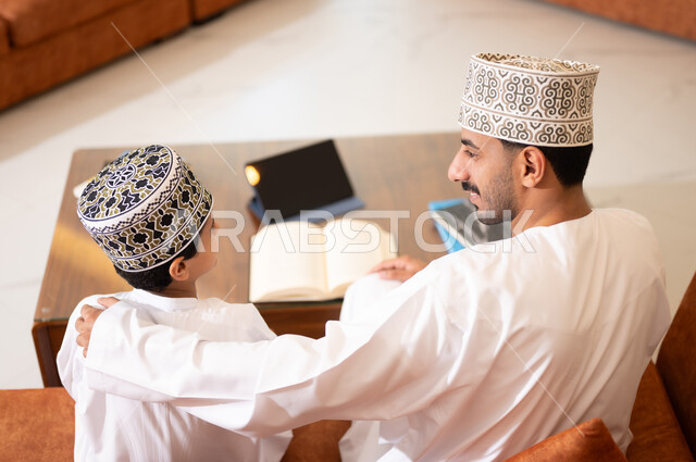 تنميه قدرات الاطفال بالتعلم، تشجيع وتطوير الاطفال بحل الواجبات المدرسيه المنزليه، بورتريه لأب عربي خليجي عماني يرتدي الدشداشه والكمه يقوم بمساعده ابنه في التعليم والقراءه، الانشطة التعليمية الالكترونيه  وانجاز الدروس والواجبات عن بعد باستخدام جهاز الايباد المحمول