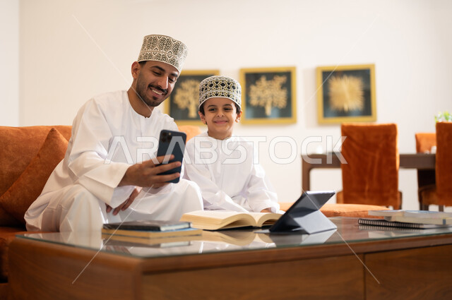 Developing children’s learning abilities, encouraging and developing children by solving home school assignments, a portrait of an Arab, Gulf, Omani father wearing a dishdasha and a sleeve, helping his son in education and reading, electronic educational activities, and completing lessons and assignments remotely using a portable iPad.