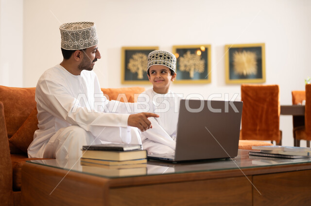 Developing children’s learning abilities, encouraging and developing children by solving home school assignments, a portrait of an Arab, Gulf, Omani father wearing a dishdasha and a sleeve, helping his son in education and reading, electronic educational activities, and completing lessons and assignments remotely using a portable iPad.