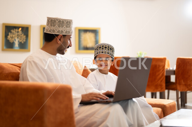 تنميه قدرات الاطفال بالتعلم، تشجيع وتطوير الاطفال بحل الواجبات المدرسيه المنزليه، بورتريه لأب عربي خليجي عماني يرتدي الدشداشه والكمه يقوم بمساعده ابنه في التعليم والقراءه، الانشطة التعليمية الالكترونيه  وانجاز الدروس والواجبات عن بعد باستخدام جهاز الايباد المحمول