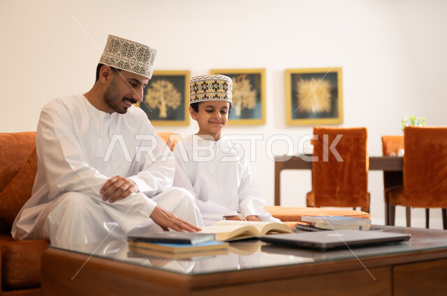 Developing children’s learning abilities, encouraging and developing children by solving home school assignments, a portrait of an Arab, Gulf, Omani father wearing a dishdasha and a sleeve, helping his son in education and reading, electronic educational activities, and completing lessons and assignments remotely using a portable iPad.