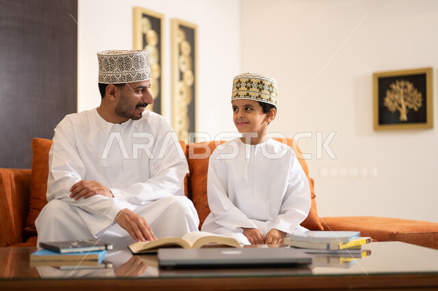 تنميه قدرات الاطفال بالتعلم، تشجيع وتطوير الاطفال بحل الواجبات المدرسيه المنزليه، بورتريه لأب عربي خليجي عماني يرتدي الدشداشه والكمه يقوم بمساعده ابنه في التعليم والقراءه، الانشطة التعليمية الالكترونيه  وانجاز الدروس والواجبات عن بعد باستخدام جهاز الايباد المحمول