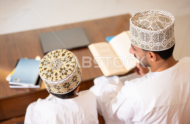 تنميه قدرات الاطفال بالتعلم، تشجيع وتطوير الاطفال بحل الواجبات المدرسيه المنزليه، بورتريه لأب عربي خليجي عماني يرتدي الدشداشه والكمه يقوم بمساعده ابنه في التعليم والقراءه، الانشطة التعليمية الالكترونيه  وانجاز الدروس والواجبات عن بعد باستخدام جهاز الايباد المحمول