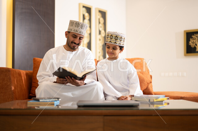 تنميه قدرات الاطفال بالتعلم، تشجيع وتطوير الاطفال بحل الواجبات المدرسيه المنزليه، بورتريه لأب عربي خليجي عماني يرتدي الدشداشه والكمه يقوم بمساعده ابنه في التعليم والقراءه، الانشطة التعليمية الالكترونيه  وانجاز الدروس والواجبات عن بعد باستخدام جهاز الايباد المحمول