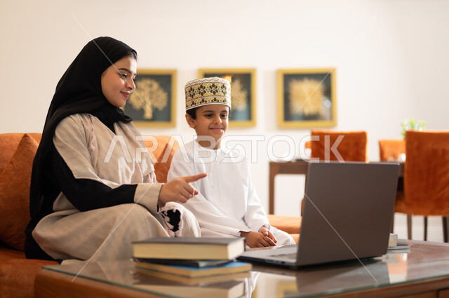 تنميه قدرات الاطفال بالتعلم، تشجيع وتطوير الاطفال بحل الواجبات المدرسيه المنزليه، بورتريه لأم عربية خليجية عمانيه محجبة تساعد طفلها في التعليم والقراءه، الانشطة التعليمية الالكترونيه  وانجاز الدروس والواجبات عن بعد باستخدام جهاز الايباد المحمول