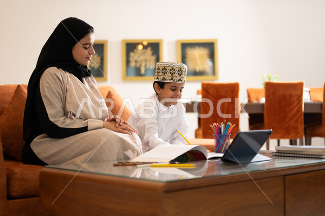 تنميه قدرات الاطفال بالتعلم، تشجيع وتطوير الاطفال بحل الواجبات المدرسيه المنزليه، بورتريه لأم عربية خليجية عمانيه محجبة تساعد طفلها في التعليم والقراءه، الانشطة التعليمية الالكترونيه  وانجاز الدروس والواجبات عن بعد باستخدام جهاز الايباد المحمول