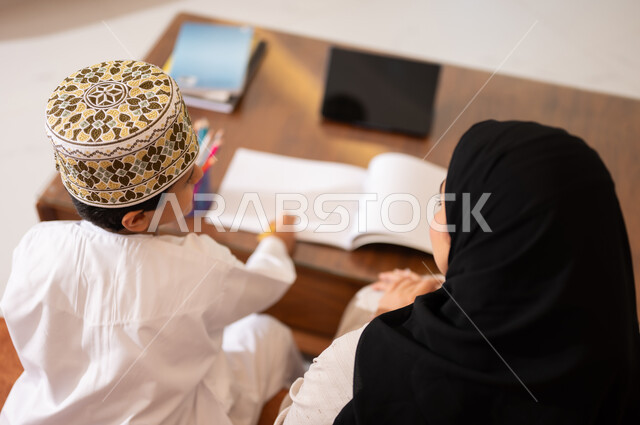 تنميه قدرات الاطفال بالتعلم، تشجيع وتطوير الاطفال بحل الواجبات المدرسيه المنزليه، بورتريه لأم عربية خليجية عمانيه محجبة تساعد طفلها في التعليم والقراءه، الانشطة التعليمية الالكترونيه  وانجاز الدروس والواجبات عن بعد باستخدام جهاز الايباد المحمول