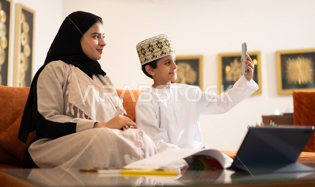 تنميه قدرات الاطفال بالتعلم، تشجيع وتطوير الاطفال بحل الواجبات المدرسيه المنزليه، بورتريه لأم عربية خليجية عمانيه محجبة تساعد طفلها في التعليم والقراءه، الانشطة التعليمية الالكترونيه  وانجاز الدروس والواجبات عن بعد باستخدام جهاز الايباد المحمول