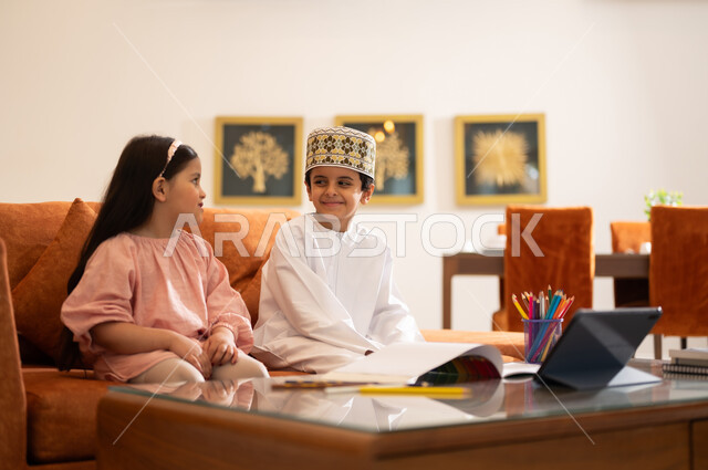 مراجعة الدروس اليومية عبر الانترنت، تطور العلم والتعليم في سلطنه عمان، بورتريه لتلميذين عربيين خليجيين عمانييين يرتدون الزي العماني التقليدي ويجلسون في غرفه المعيشه امام جهاز التابلت المحمول يقومون بحل الواجبات المدرسيه وتلقي المحاضرات المدرسيه عن بعد